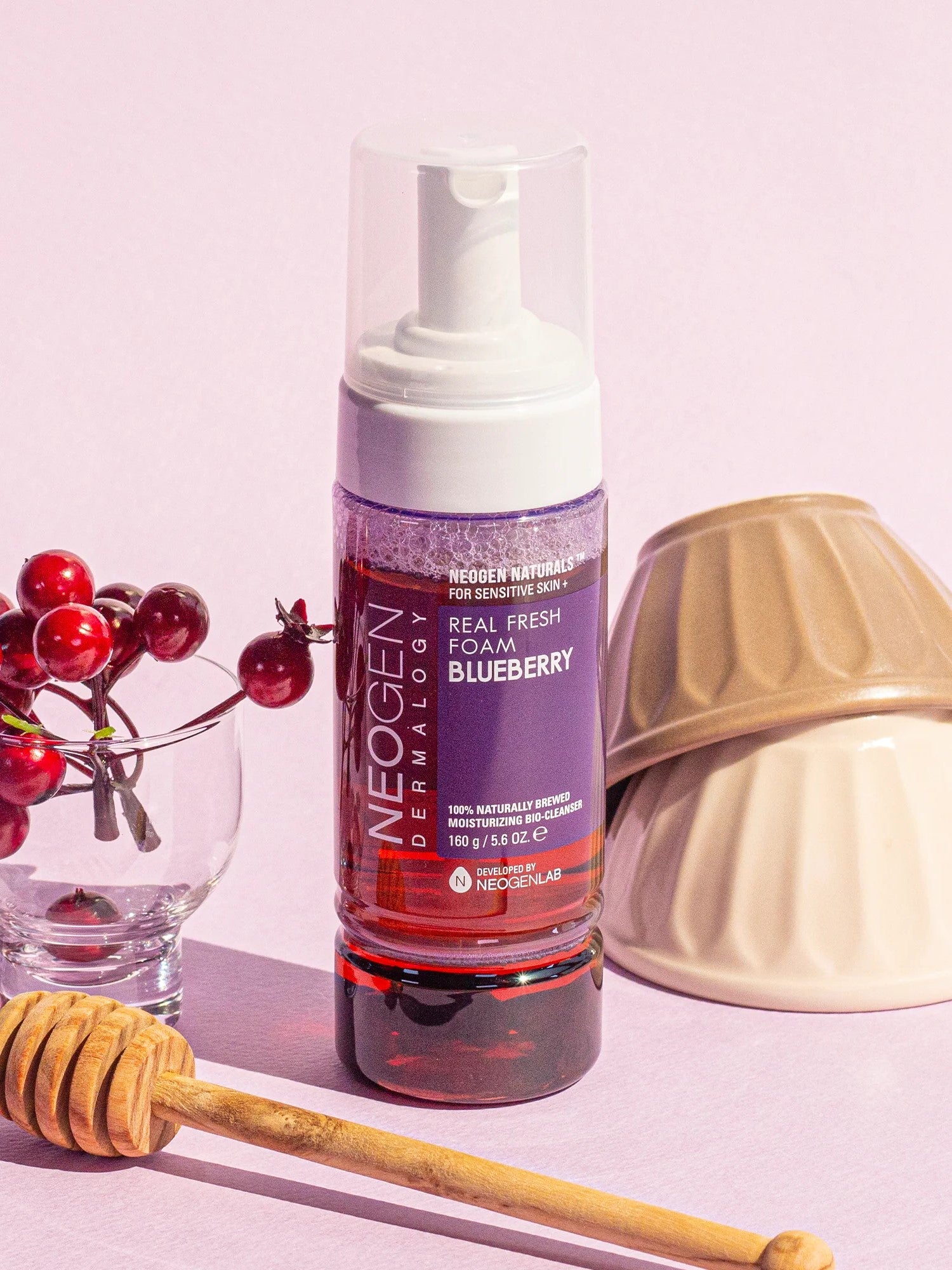 Neogen Real Fresh Foam Blueberry Cleanser bottle displayed alongside fresh blueberries and a honey dipper on a pastel pink backdrop, emphasizing its antioxidant-rich formula and gentle cleansing benefits.
