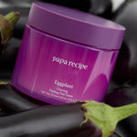A purple jar labeled 'Papa Recipe Eggplant Clearing Peeling Pad Toner,' surrounded by fresh eggplants on a dark background, emphasizing natural ingredients.