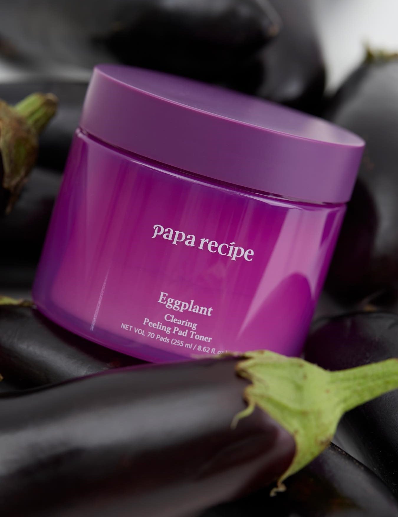 A purple jar labeled 'Papa Recipe Eggplant Clearing Peeling Pad Toner,' surrounded by fresh eggplants on a dark background, emphasizing natural ingredients.
