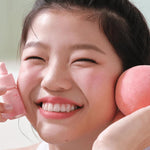 Model holding Peripera INK AIRY VELVET 23 Peaches Collection lip tint in one hand and a peach-colored beauty sponge in the other, against a light background.
