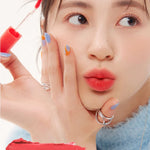 A model wearing Romand Blur Fudge Tint in the shade Coral Jublee, showcasing the soft matte texture on her lips.