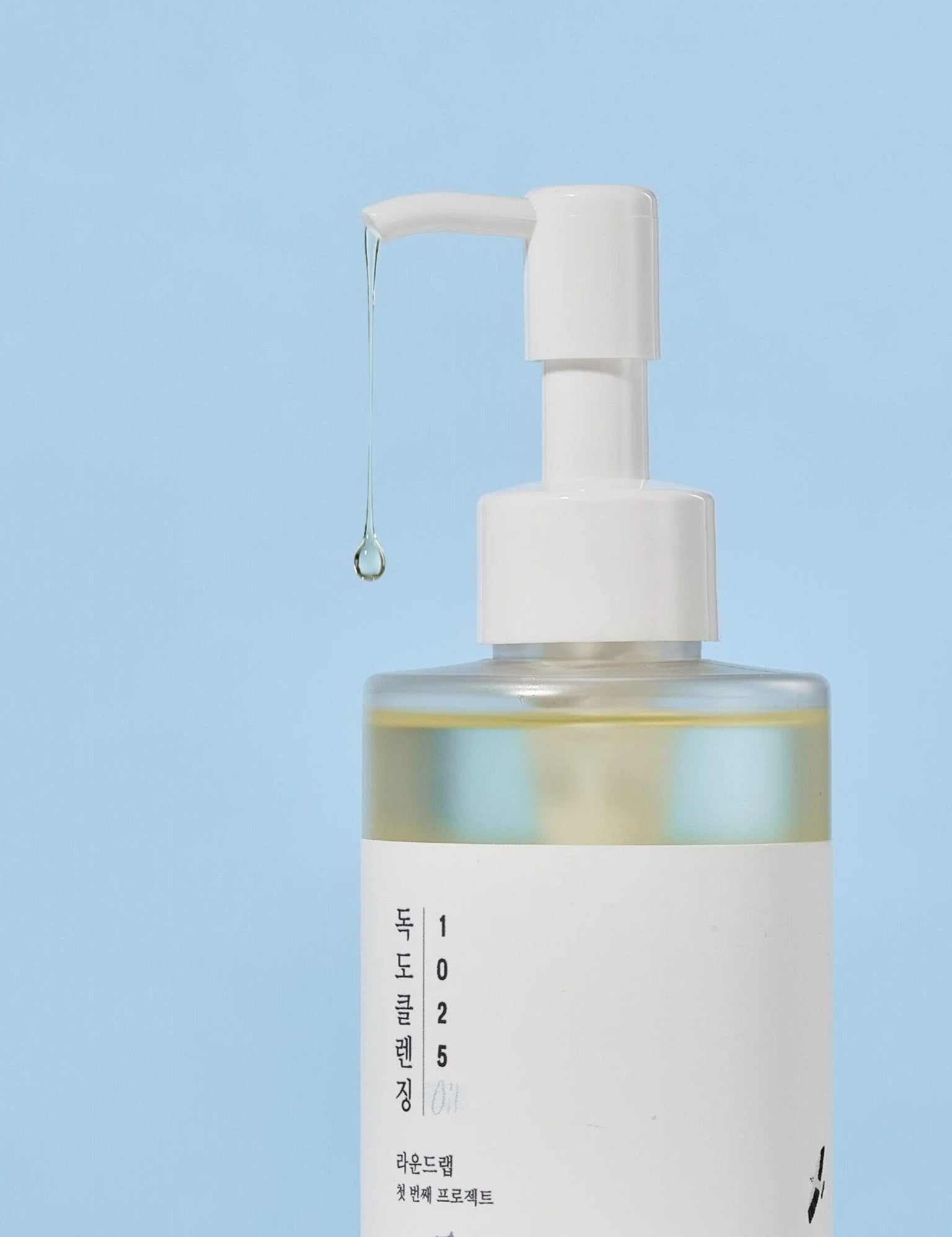Round Lab 1025 Dokdo Cleansing Oil with liquid dripping from the nozzle, set against a soft blue background.