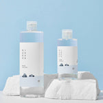 Two bottles of Round Lab 1025 Dokdo Toner with minimalist labels, arranged on white rocks against a soft blue background.