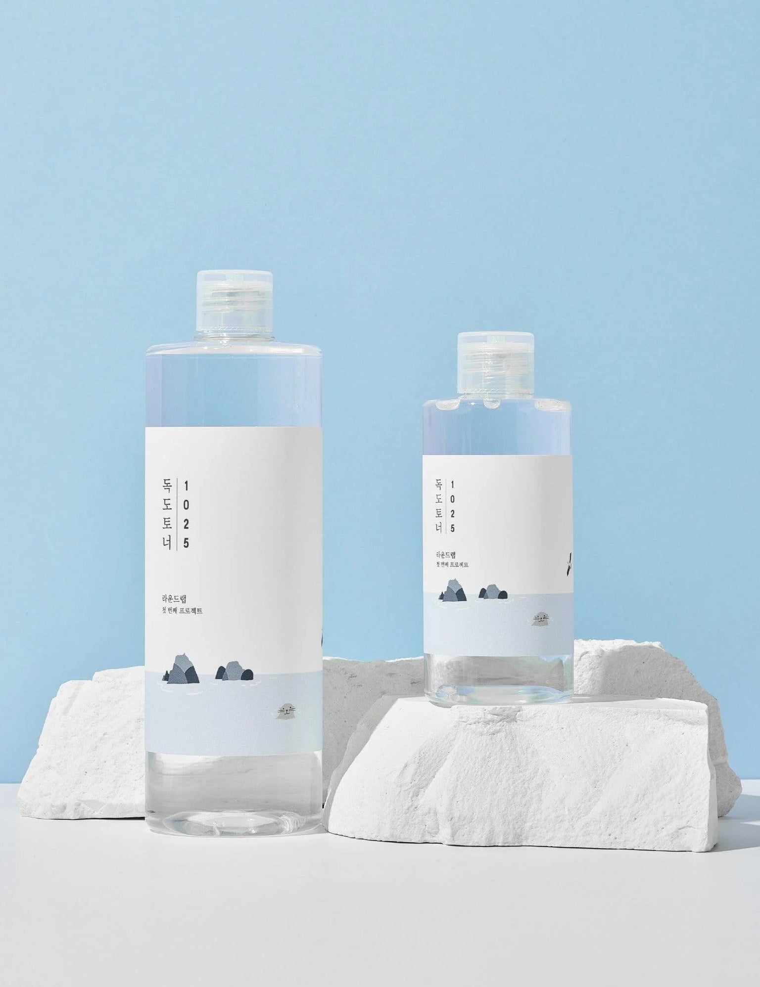 Two bottles of Round Lab 1025 Dokdo Toner with minimalist labels, arranged on white rocks against a soft blue background.
