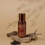 A bottle of Tonymoly The Black Tea London Classic Serum resting on stones, with loose tea leaves and twigs to highlight its natural ingredients, set against a beige backdrop.
