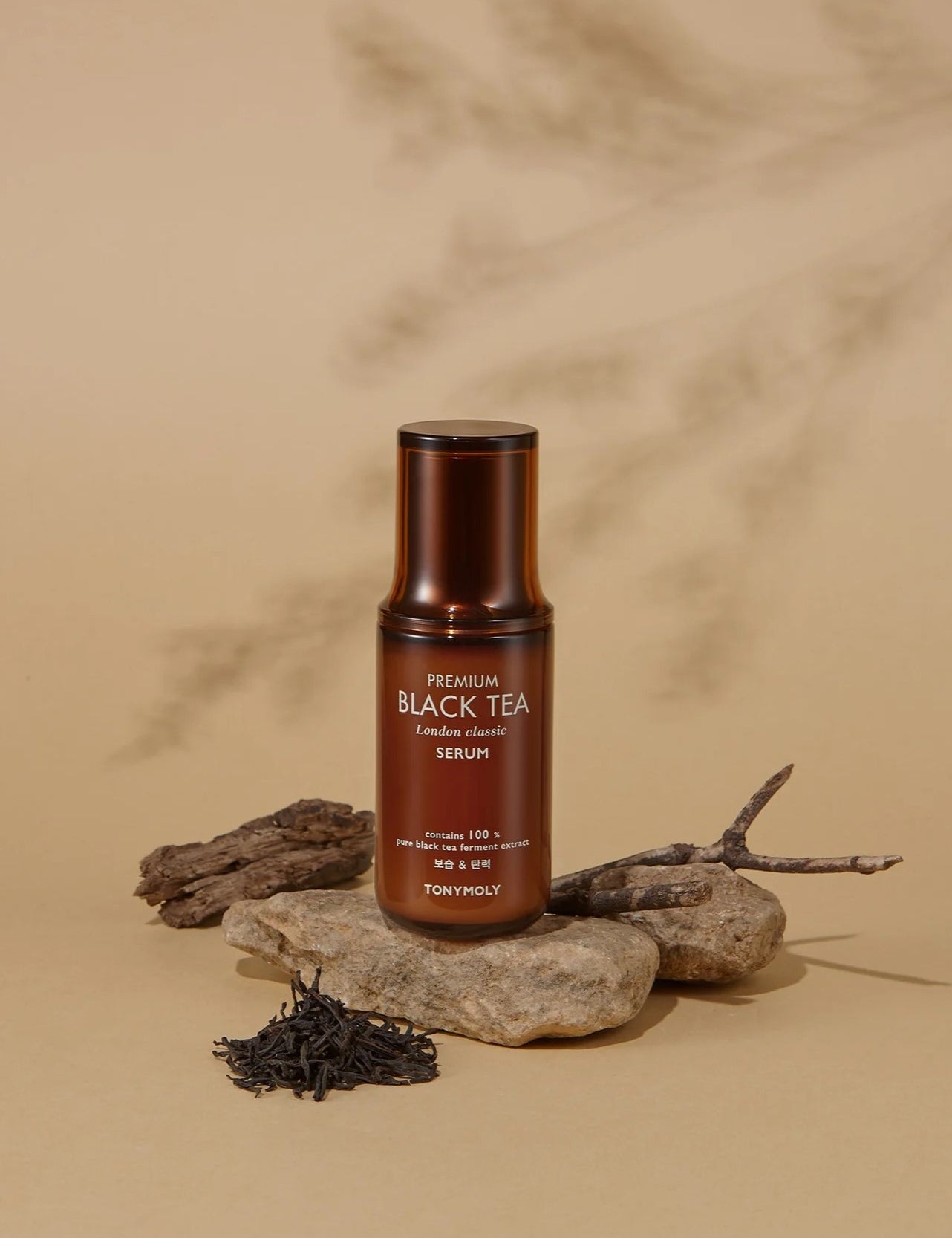 A bottle of Tonymoly The Black Tea London Classic Serum resting on stones, with loose tea leaves and twigs to highlight its natural ingredients, set against a beige backdrop.