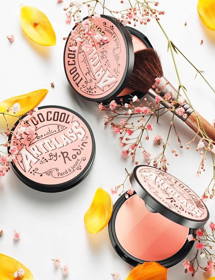 Too Cool for School Artclass By Rodin Blush in De Peche shade, surrounded by soft flowers and petals, with a makeup brush for effortless peach-toned application.