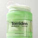 Green jar of Torriden Balanceful Toner Pad next to a stack of its white pads, with the label highlighting Centella Asiatica Extract for soothing and hydration.
