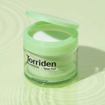 Transparent jar of Torriden Balanceful Toner Pad with a green lid, placed on a soft green rippled surface, showcasing its fresh and minimalist aesthetic.