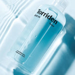 Clear toner bottle labeled 'Torriden Dive In' with 'Low Molecular Hyaluronic Acid,' sitting on wavy blue water background.