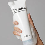 Hand holding a foamy tube of Torriden Dive In Cleansing Foam, with visible texture and labeled 'Low Molecular Hyaluronic Acid,' against a neutral background.