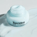 Jar of Torriden Dive In Low Molecular Hyaluronic Acid Soothing Cream on a reflective surface, surrounded by a creamy, light blue product spill.