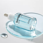 Clear blue serum bottle with a dropper, labeled 'Torriden Dive In,' resting on a glass plate surrounded by droplets of its lightweight texture.