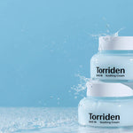 Two jars of Torriden Dive In Soothing Cream on a reflective surface, splashed with droplets against a soft blue background.
