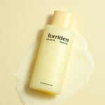 A bottle of Torriden Solid IN Essence with a pale yellow label sitting atop its creamy yet lightweight essence pool against a soft yellow background.