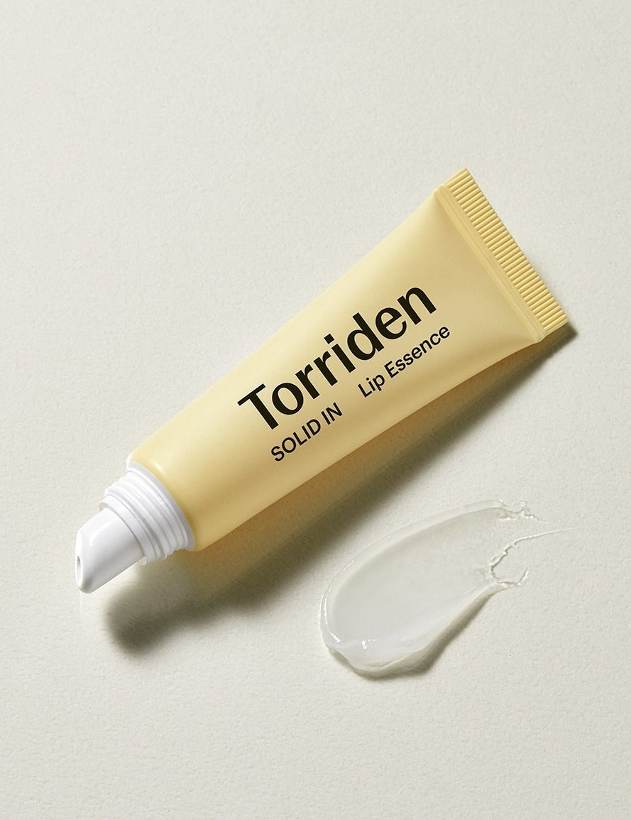 A yellow tube of Torriden Solid In Lip Essence with a white nozzle, next to a glossy droplet of the lip essence on a textured surface.