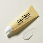 A yellow tube of Torriden Solid In Lip Essence with a white nozzle, next to a glossy droplet of the lip essence on a textured surface.