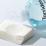 Bottle of Torriden toner next to stacked cotton pads, with a small amount of toner dispensed onto one pad.