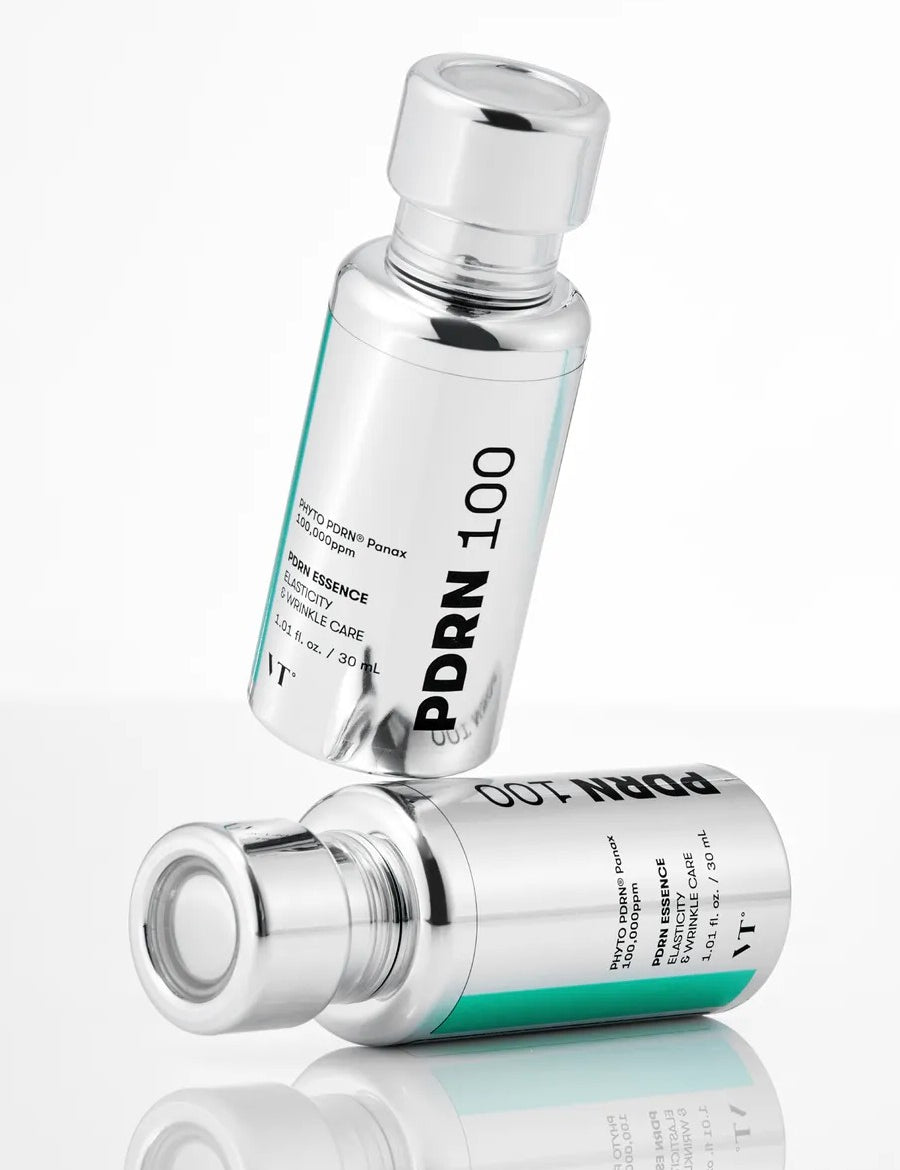 Two silver bottles of VT Cosmetics PDRN Essence 100, designed for elasticity and wrinkle care, displayed on a reflective surface.
